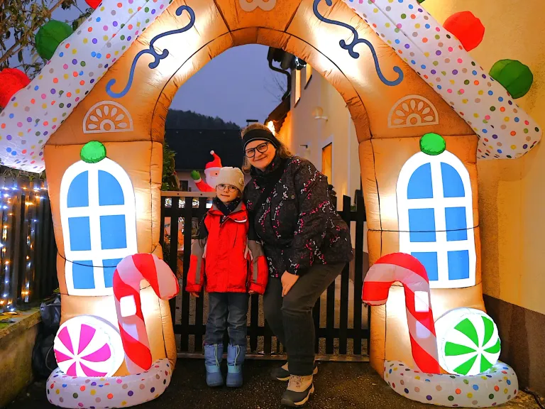 Eine Frau und ein Kind stehen vor dem Eingang eines Lebkuchenhauses. Die Frau lächelt und das Kind trägt einen Weihnachtsmannhut.