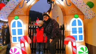 A woman and a child are standing in front of a gingerbread house entrance. The woman is smiling, and the child is wearing a Santa hat.