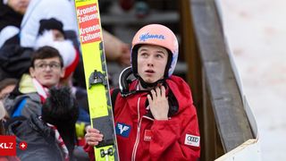 Ein Skifahrer mit einem rosa Helm, auf dem das Wort Manner steht, hält Skier in der linken Hand. Er steht auf einer Skisprungschanze und sieht konzentriert auf etwas. Dahinter ist ein verschwommenes Bild einer Menge, die das Event beobachtet.
