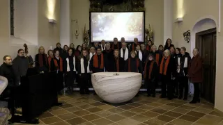 Ein Chor steht um einen großen Marmorbrunnen in einer Kirche, mit einem Leinwandprojektor dahinter. Sie tragen rote Schals und schwarze Kleidung.