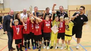 Eine Gruppe junger Fußballspieler feiert mit einem Pokal und Medaillen in einer Turnhalle, umgeben von Erwachsenen und einem Schiedsrichter.