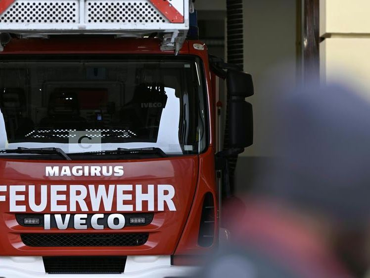 Ein roter Feuerwehrwagen mit weißen Buchstaben 'MAGIRUS FEUERWEHR IVECO', geparkt in einer Garage mit einer verschwommenen Figur im Hintergrund.