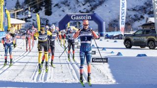 Langläufer treten bei einem Langlaufrennen auf einer verschneiten Strecke an, mit Bannern und Fahnen im Hintergrund.