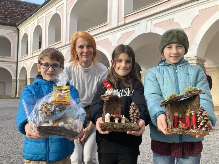 Drei Kinder und eine Frau halten handgefertigte Krippen vor einem Gebäude mit Bögen. Die Kinder lächeln und die Frau schaut sie an. Die Kinder halten ihre Kreationen, während die Frau eine kleine Tasche hält.