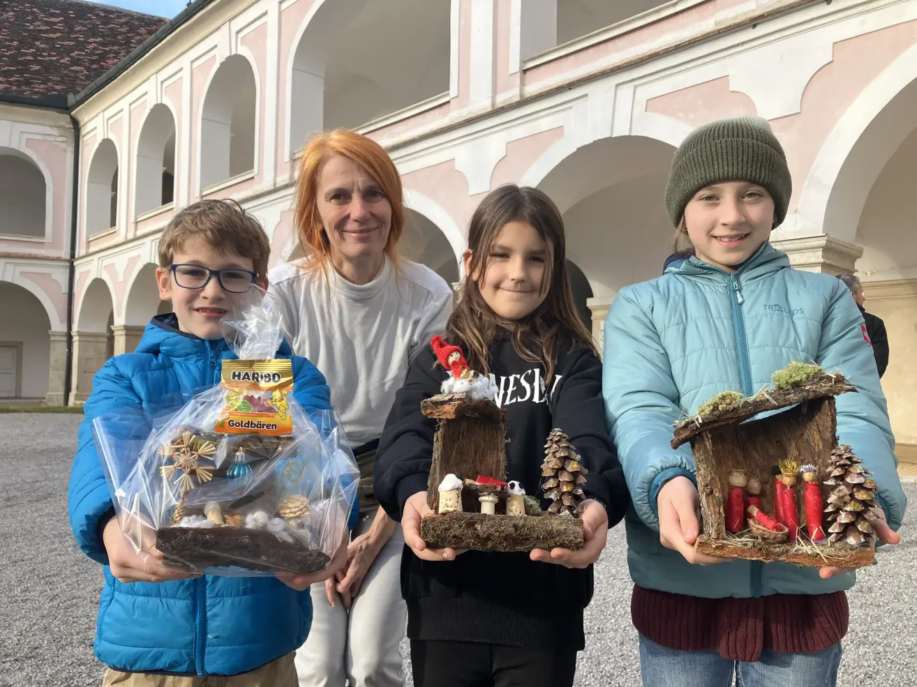Drei Kinder und eine Frau halten handgefertigte Krippen vor einem Gebäude mit Bögen. Die Kinder lächeln und die Frau schaut sie an. Die Kinder halten ihre Kreationen, während die Frau eine kleine Tasche hält.