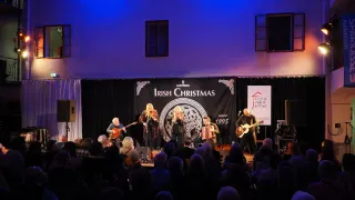 Eine Live-Performance auf der Bühne mit Musikern, die Gitarren, Akkordeons und andere Instrumente spielen. Das Publikum sitzt vor der Bühne, und ein Banner mit dem Text 'Irish Christmas' hängt hinter den Künstlern.