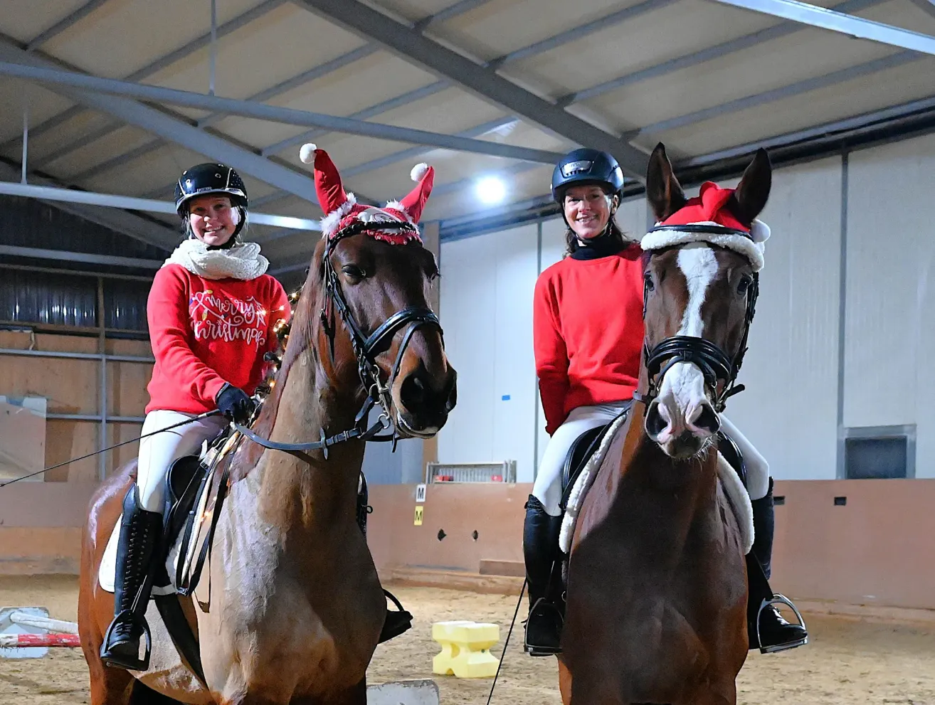 Two women riding horses in a stable. Both women and horses are wearing Christmas hats. The woman on the left is smiling, and the woman on the right is posing.