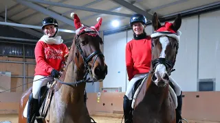 Two women riding horses in a stable. Both women and horses are wearing Christmas hats. The woman on the left is smiling, and the woman on the right is posing.