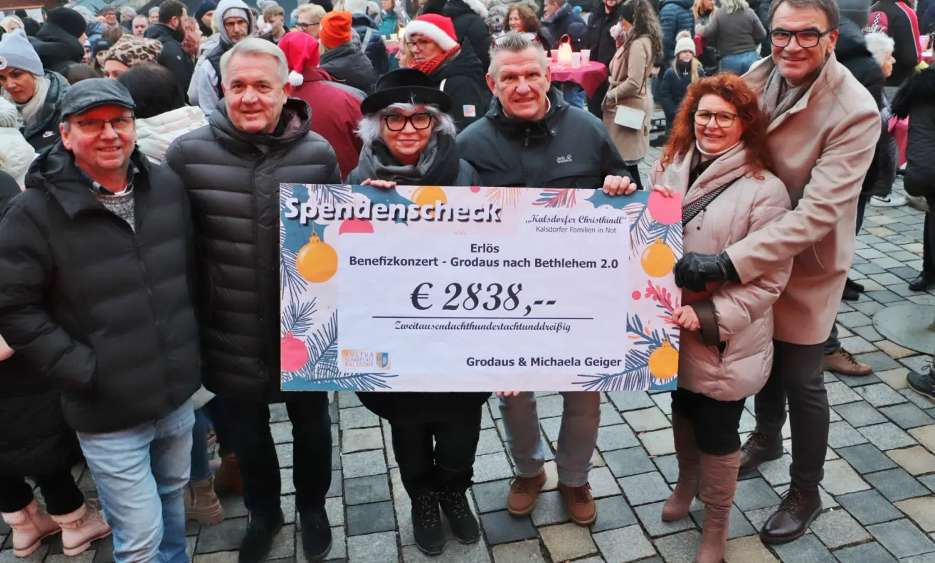 Vier Menschen stehen zusammen und halten einen großen Scheck für 2838 Euro. Sie befinden sich in einer festlichen Umgebung mit Winterkleidung, lächeln und sind von einer Menge umgeben.