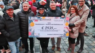 Vier Menschen stehen zusammen und halten einen großen Scheck für 2838 Euro. Sie befinden sich in einer festlichen Umgebung mit Winterkleidung, lächeln und sind von einer Menge umgeben.
