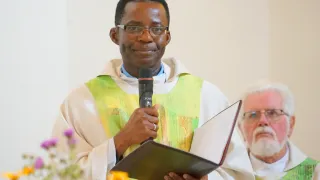Ein Priester in grüner Amtstracht hält ein Mikrofon und ein Buch und spricht während eines Gottesdienstes. Ein weiterer Priester steht im Hintergrund.
