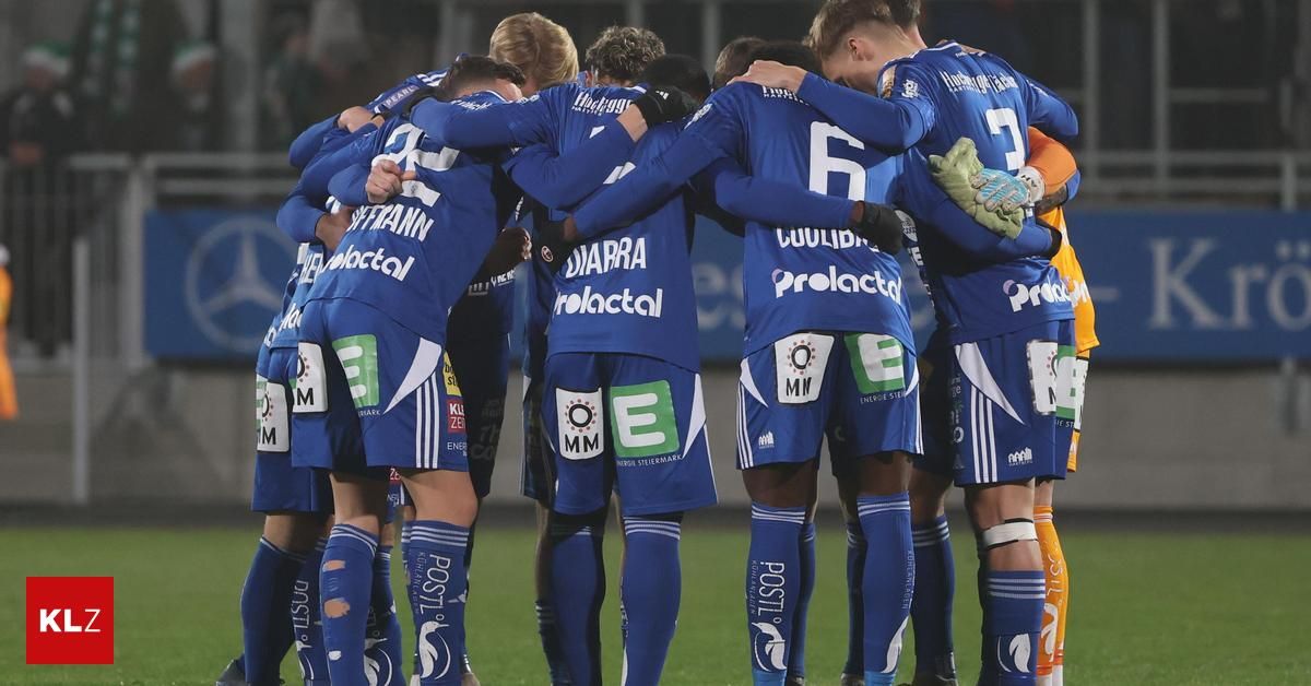 Eine Gruppe von Fußballspielern in blauen Trikots drängt sich auf dem Spielfeld zusammen. Jeder Spieler trägt eine Nummer auf dem Rücken und scheint sich auf eine Strategie zu konzentrieren.