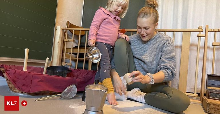 Eine Mutter und ihre kleine Tochter sitzen auf dem Boden und beschäftigen sich mit einer spielerischen Aktivität. Die Mutter hält ein kleines Objekt, möglicherweise ein Spielzeug, während das Mädchen lächelt zuschaut. In der Nähe sind Küchenutensilien und ein Korb zu sehen.