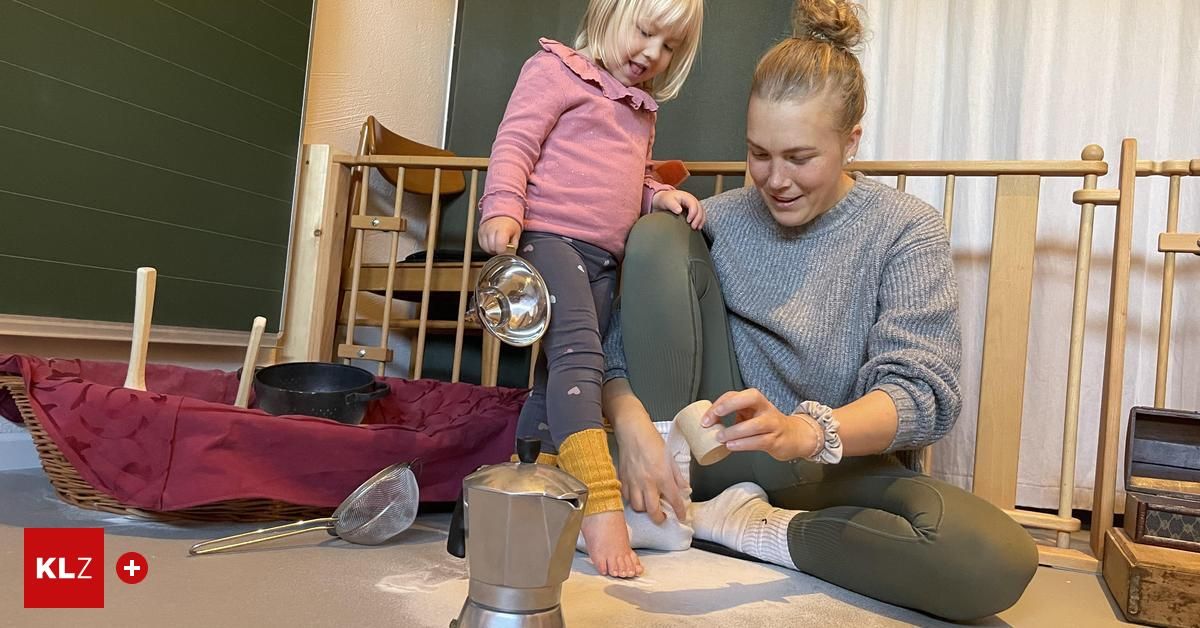 Eine Mutter und ihre kleine Tochter sitzen auf dem Boden und beschäftigen sich mit einer spielerischen Aktivität. Die Mutter hält ein kleines Objekt, möglicherweise ein Spielzeug, während das Mädchen lächelt zuschaut. In der Nähe sind Küchenutensilien und ein Korb zu sehen.