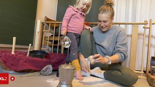 Eine Mutter und ihre kleine Tochter sitzen auf dem Boden und beschäftigen sich mit einer spielerischen Aktivität. Die Mutter hält ein kleines Objekt, möglicherweise ein Spielzeug, während das Mädchen lächelt zuschaut. In der Nähe sind Küchenutensilien und ein Korb zu sehen.