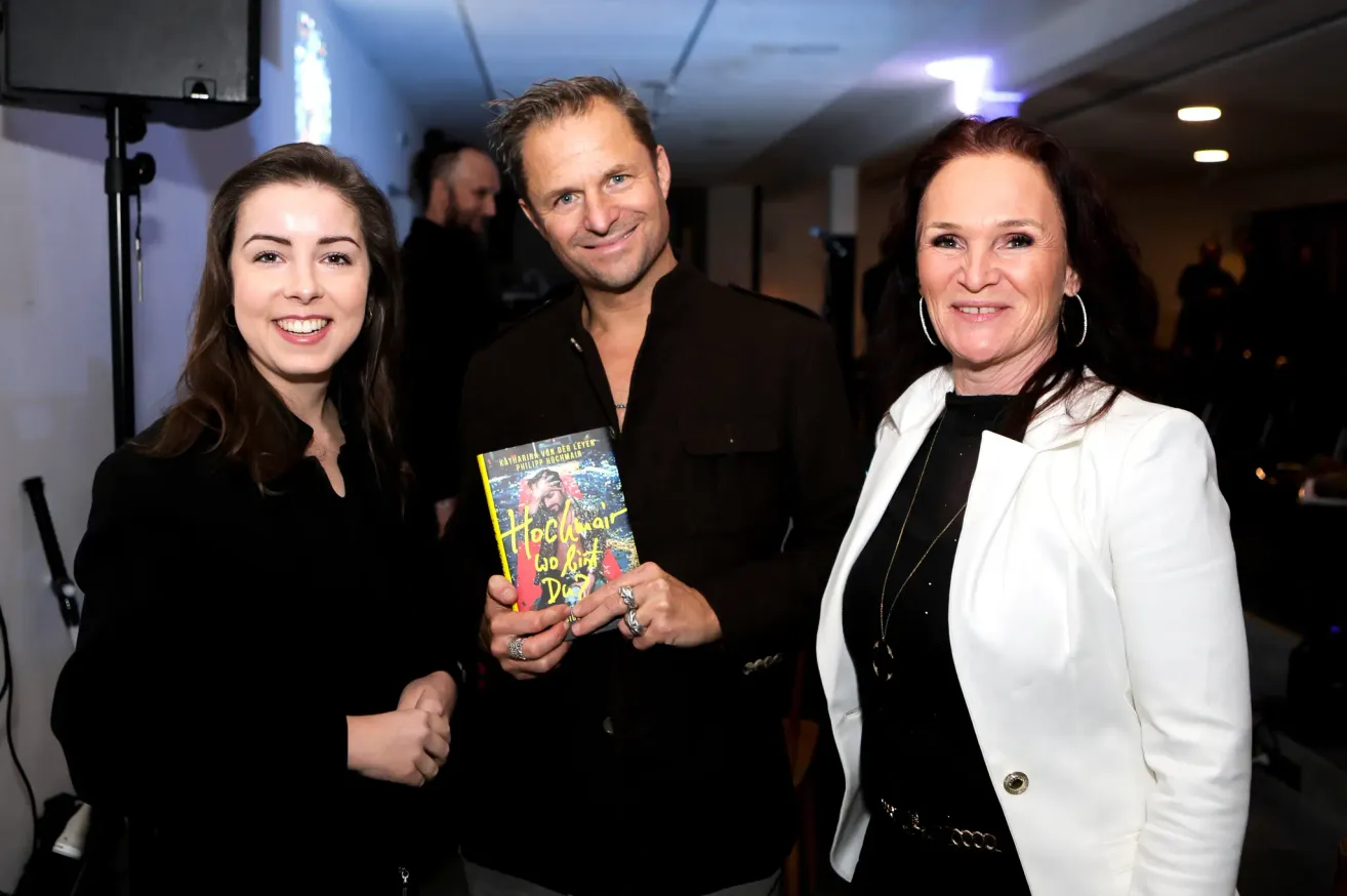 Drei Personen lächeln für ein Foto. Ein Mann in der Mitte hält ein Buch mit dem Titel 'Hochwasser'. Die Frau links trägt ein schwarzes Shirt und Ohrringe. Die Frau rechts trägt eine weiße Jacke und eine Halskette. Hinter ihnen stehen Personen und eine blaue Lichtquelle.