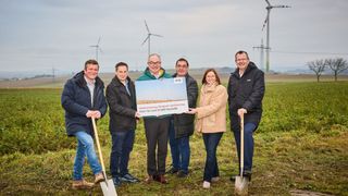 A group of six individuals, smiling and holding shovels, stand in a field with wind turbines in the background. They are holding a large blue sign with white text.