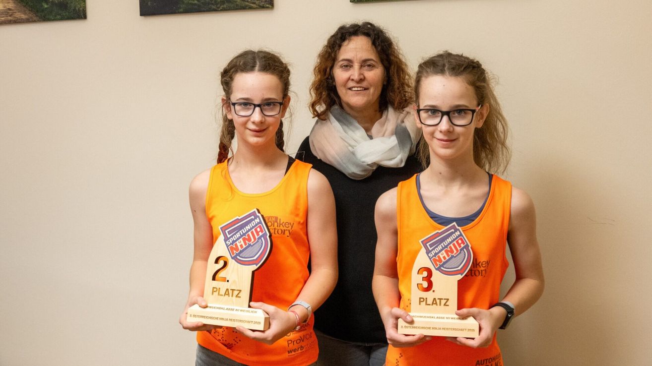 Drei Menschen stehen zusammen; zwei Mädchen halten Pokale für den zweiten und dritten Platz, und eine Frau steht lächelnd hinter ihnen.