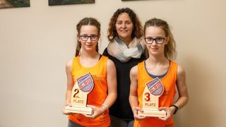 Drei Menschen stehen zusammen; zwei Mädchen halten Pokale für den zweiten und dritten Platz, und eine Frau steht lächelnd hinter ihnen.