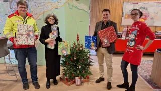 Vier Personen stehen um einen kleinen Weihnachtsbaum und halten Geschenke in den Händen. Sie befinden sich in einem Raum mit Holzwänden und einer großen Karte an einer Wand.