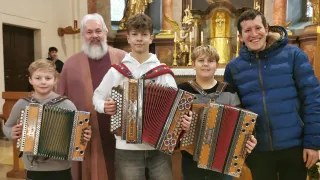 Drei Jungen mit einem älteren Mann posieren mit Akkordeons in einer Kirchenumgebung. Der Junge in der Mitte hält ein großes Akkordeon, während die anderen kleinere halten. Der Hintergrund zeigt goldene Statuen und Kerzen.