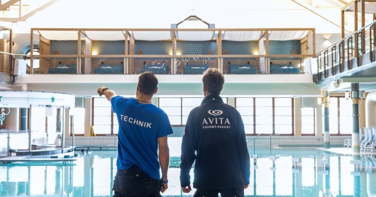 Zwei Männer in Uniformen stehen neben einem Hallenbad. Einer trägt ein blaues Shirt mit Technik, der andere ein schwarzes Jacket mit AVITA ThermeResort.