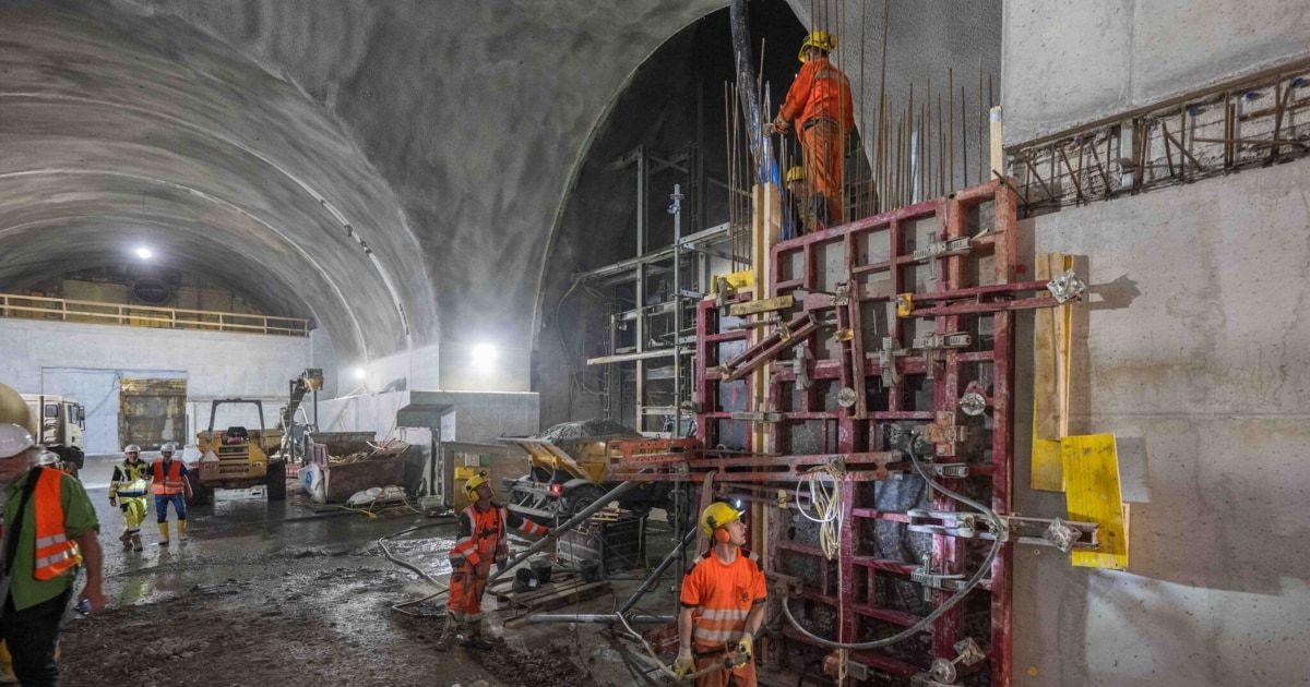 Bauarbeiter in orange Sicherheitsanzügen und Helmen arbeiten an einem Tunnelprojekt, umgeben von Maschinen und Baumaterialien.