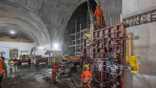 Bauarbeiter in orange Sicherheitsanzügen und Helmen arbeiten an einem Tunnelprojekt, umgeben von Maschinen und Baumaterialien.