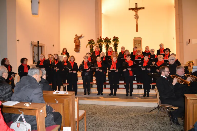 Ein Chor von Frauen in roten Schals und schwarzen Outfits steht auf den Stufen einer Kirche und singt. Ein Mann und ein Kreuz sind hinter ihnen.