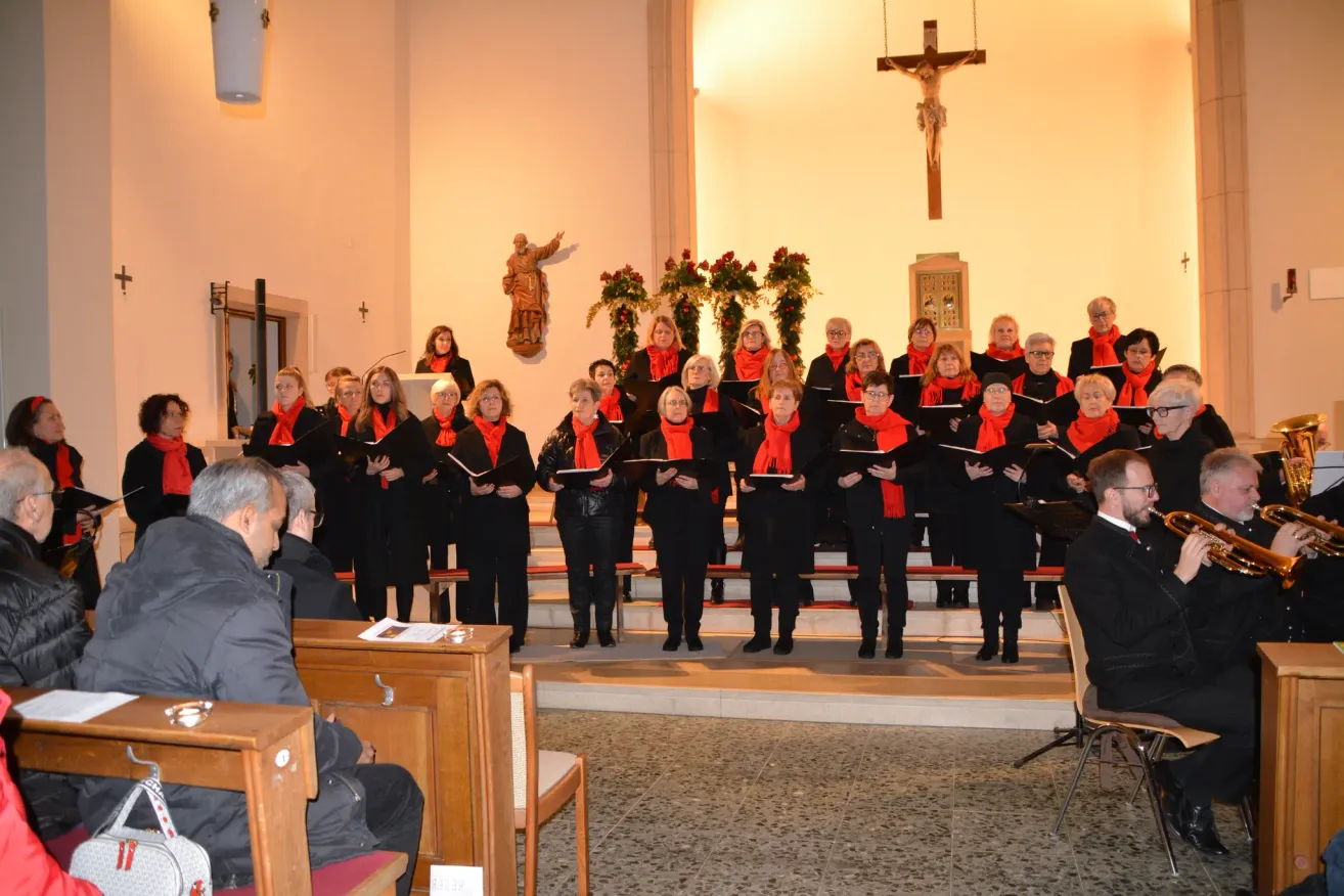 Ein Chor von Frauen in roten Schals und schwarzen Outfits steht auf den Stufen einer Kirche und singt. Ein Mann und ein Kreuz sind hinter ihnen.