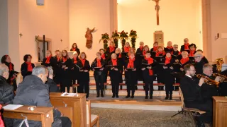 Ein Chor von Frauen in roten Schals und schwarzen Outfits steht auf den Stufen einer Kirche und singt. Ein Mann und ein Kreuz sind hinter ihnen.