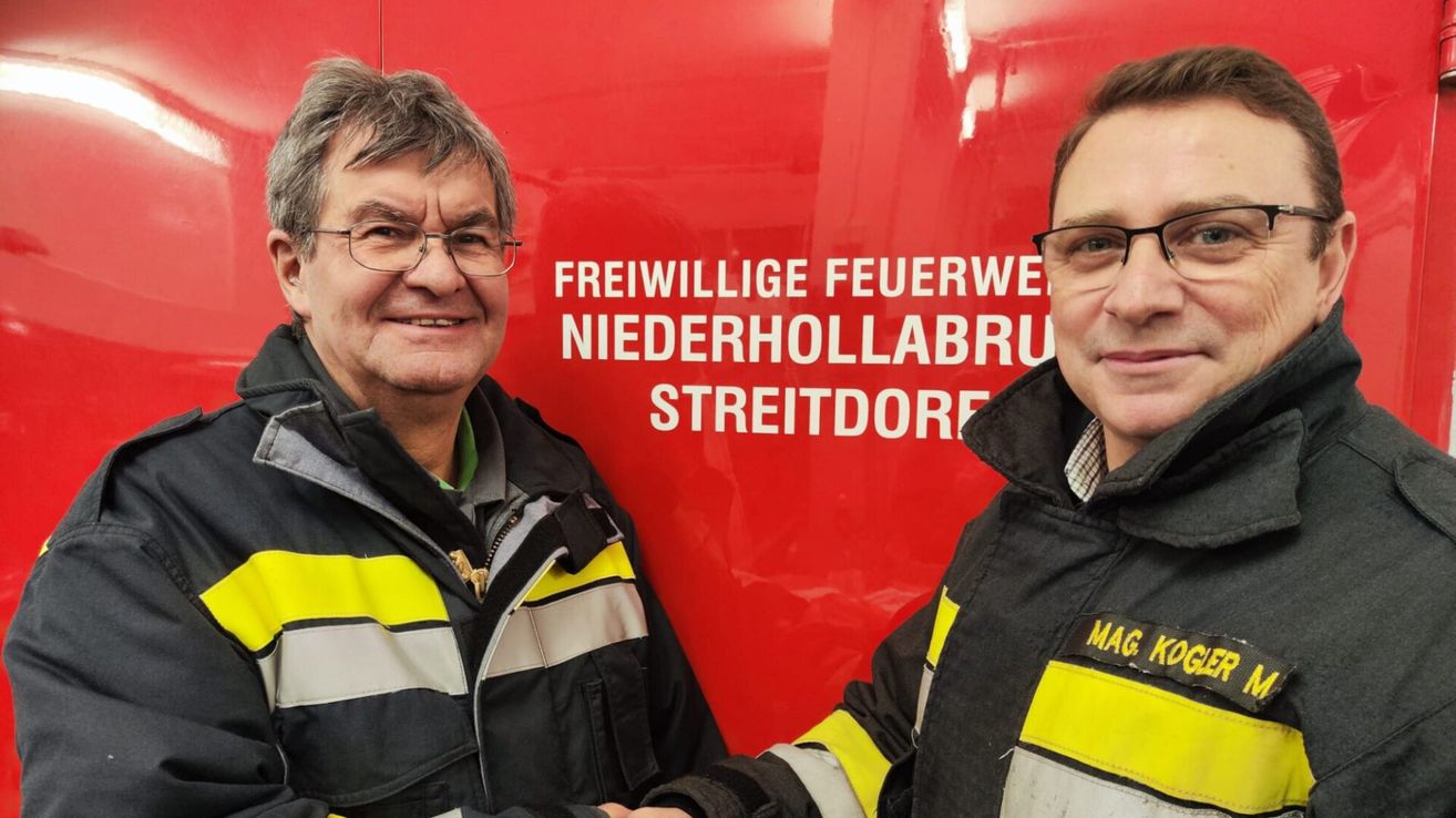 Zwei Männer in Feuerwehruniformen lächeln und schütteln Hände vor einer roten Wand. Die Wand hat den Text Freiwillige Feuerwehr Niederhollabrue Streitdorfe.