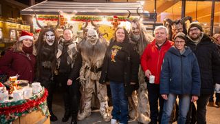 Five people are posing for a photo in a festive setting, with two wearing horned masks and others in costumes. A woman in a black jacket smiles with a cup in her hand. Behind them, a Christmas display is visible.