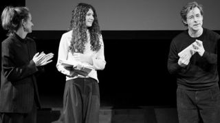 Schwarz-Weiß-Foto einer Frau, die auf einer Bühne steht, ein Buch in der Hand hält und lächelt.