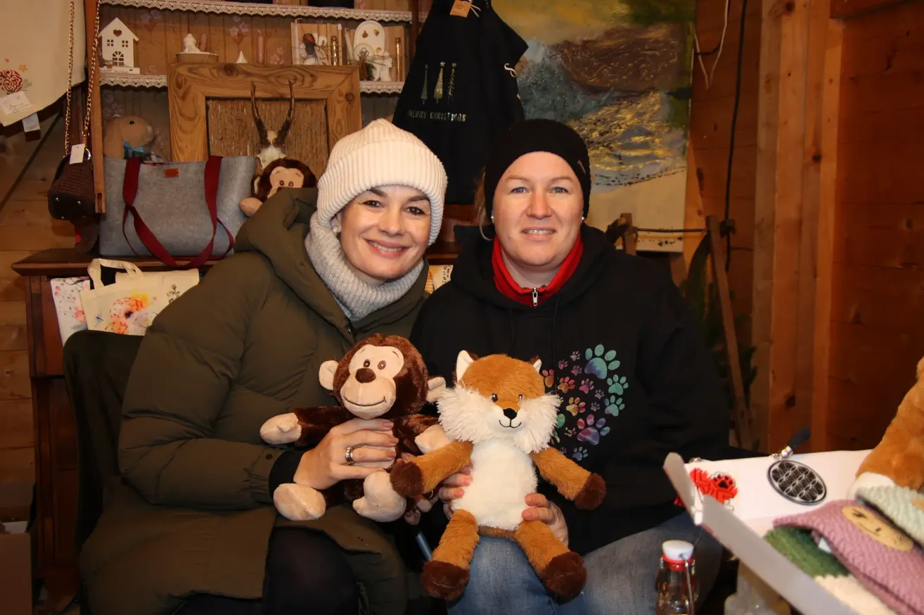 Zwei Frauen lächeln und posieren für ein Foto mit Kuscheltieren in den Händen. Eine hält einen Affen und die andere einen Fuchs. Hinter ihnen zeigt eine Wand ein Gemälde und verschiedene Gegenstände, einschließlich Handtaschen und Dekorationsartikel.