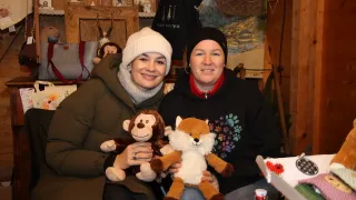 Zwei Frauen lächeln und posieren für ein Foto mit Kuscheltieren in den Händen. Eine hält einen Affen und die andere einen Fuchs. Hinter ihnen zeigt eine Wand ein Gemälde und verschiedene Gegenstände, einschließlich Handtaschen und Dekorationsartikel.