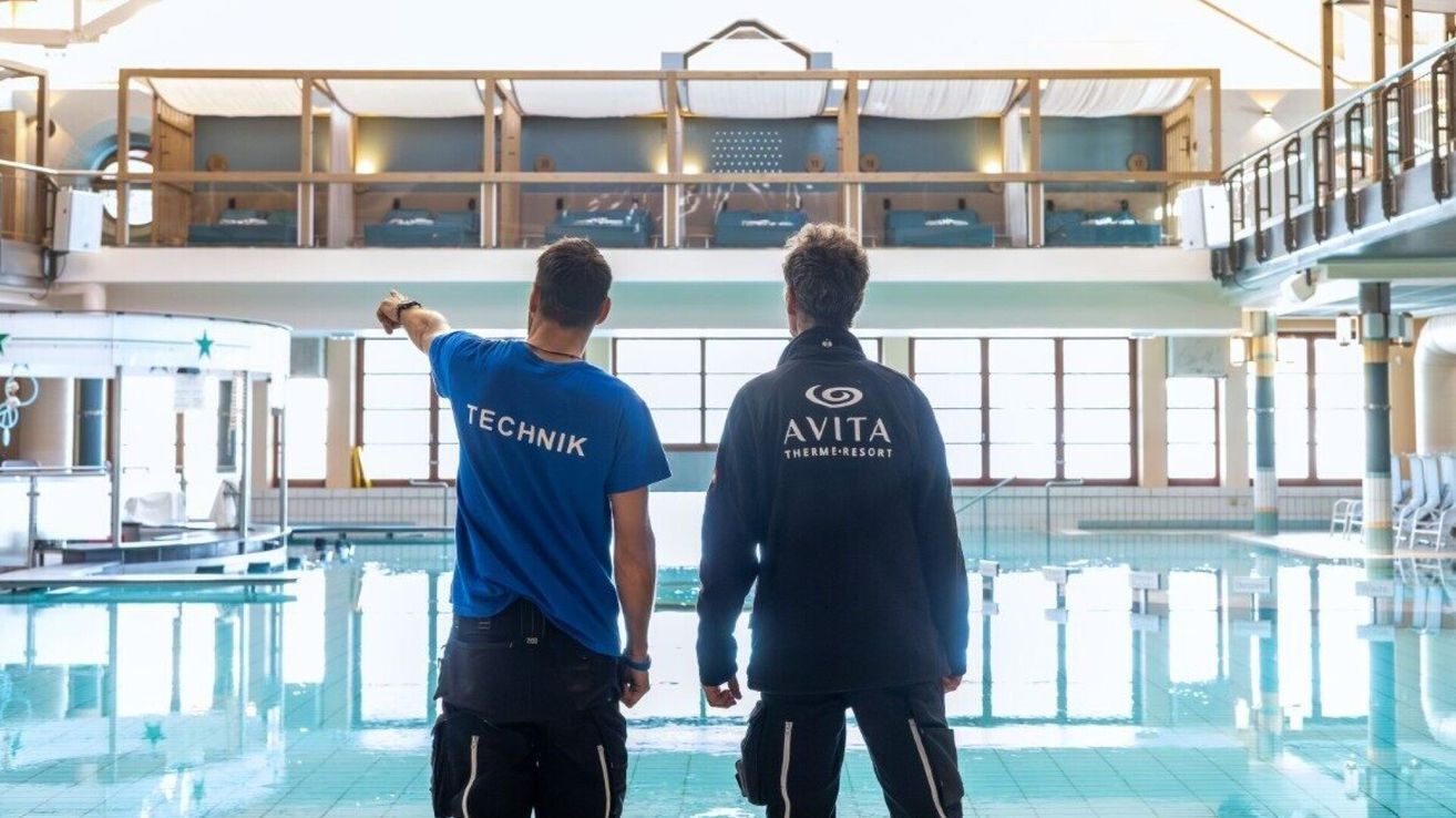Zwei Männer stehen neben einem Hallenbad, einer trägt ein blaues Technik-Shirt und der andere ein schwarzes Avita Therme-Resort-Jacke.