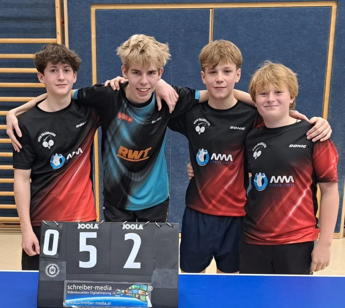 Four boys in sports uniforms pose for a photo in a gym, smiling. They stand next to a ping pong table with a scoreboard showing a score of 5 to 2.