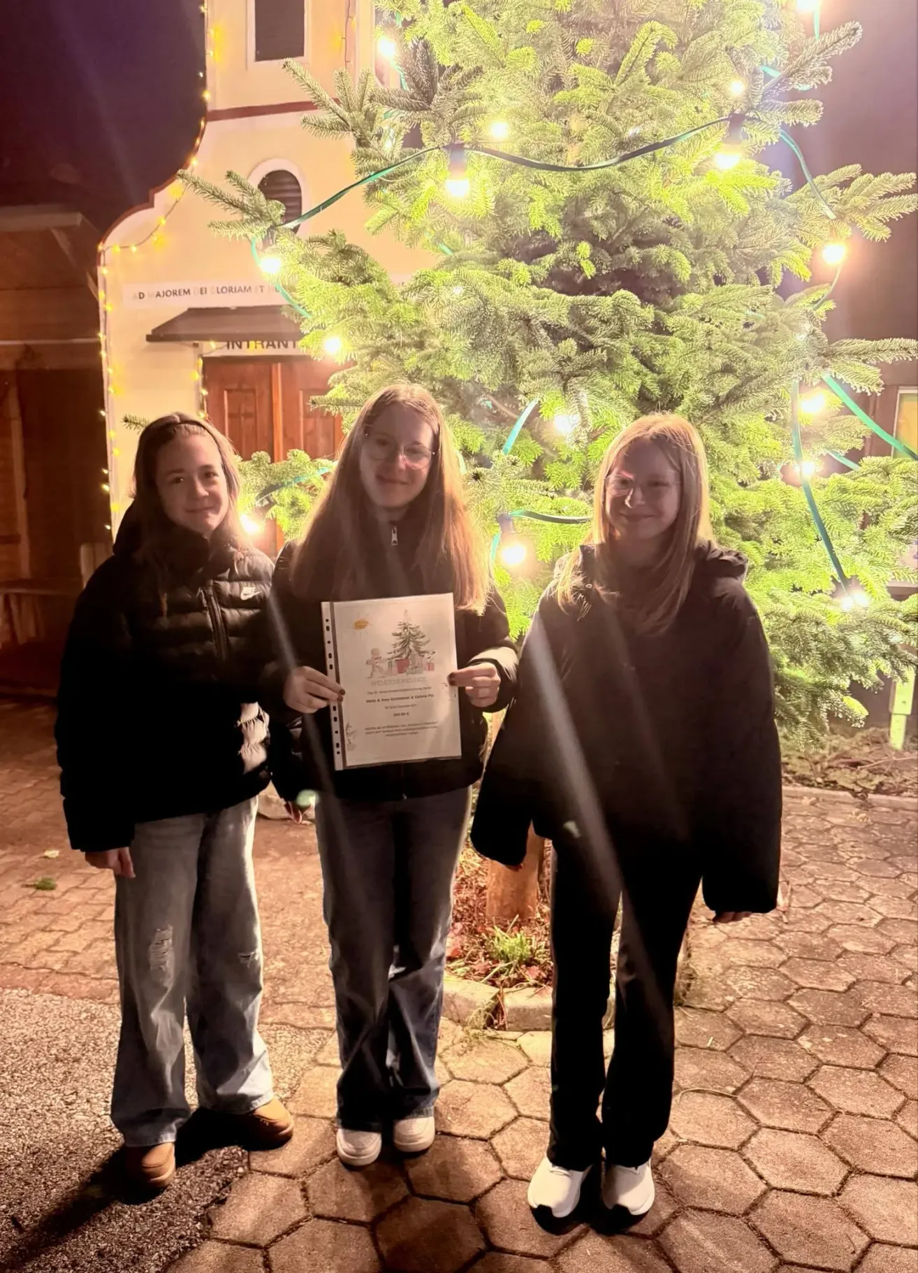 Drei junge Frauen stehen lächelnd vor einem beleuchteten Weihnachtsbaum. Die Frau in der Mitte hält ein Zertifikat. Hinter ihnen steht ein Holzhaus mit einem Schild und einem Holzzaun.