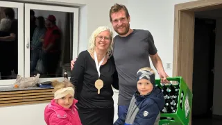 A family with two kids and a woman wearing glasses and a medal stands near a green crate filled with bottles. The man has a beard and smiles. They are in a room with white walls and a window.
