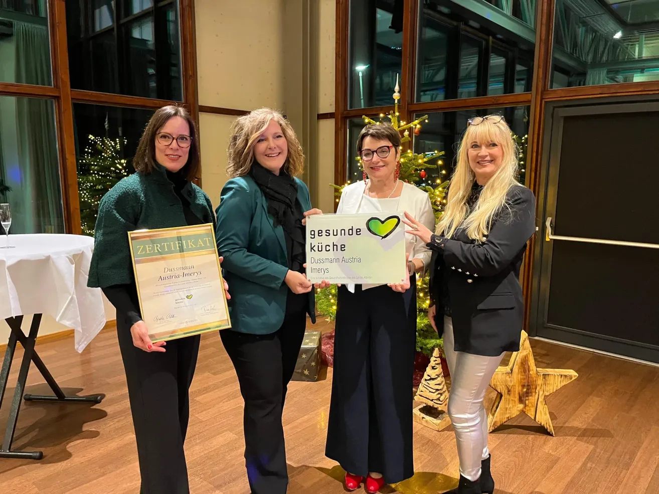 Vier Frauen stehen zusammen und posieren für ein Foto. Sie halten Zertifikate und ein Schild mit der Aufschrift 'gesunde Küche Dussmann Austria'. Sie lächeln und scheinen zu feiern. Sie sind drinnen mit einem Weihnachtsbaum hinter ihnen.