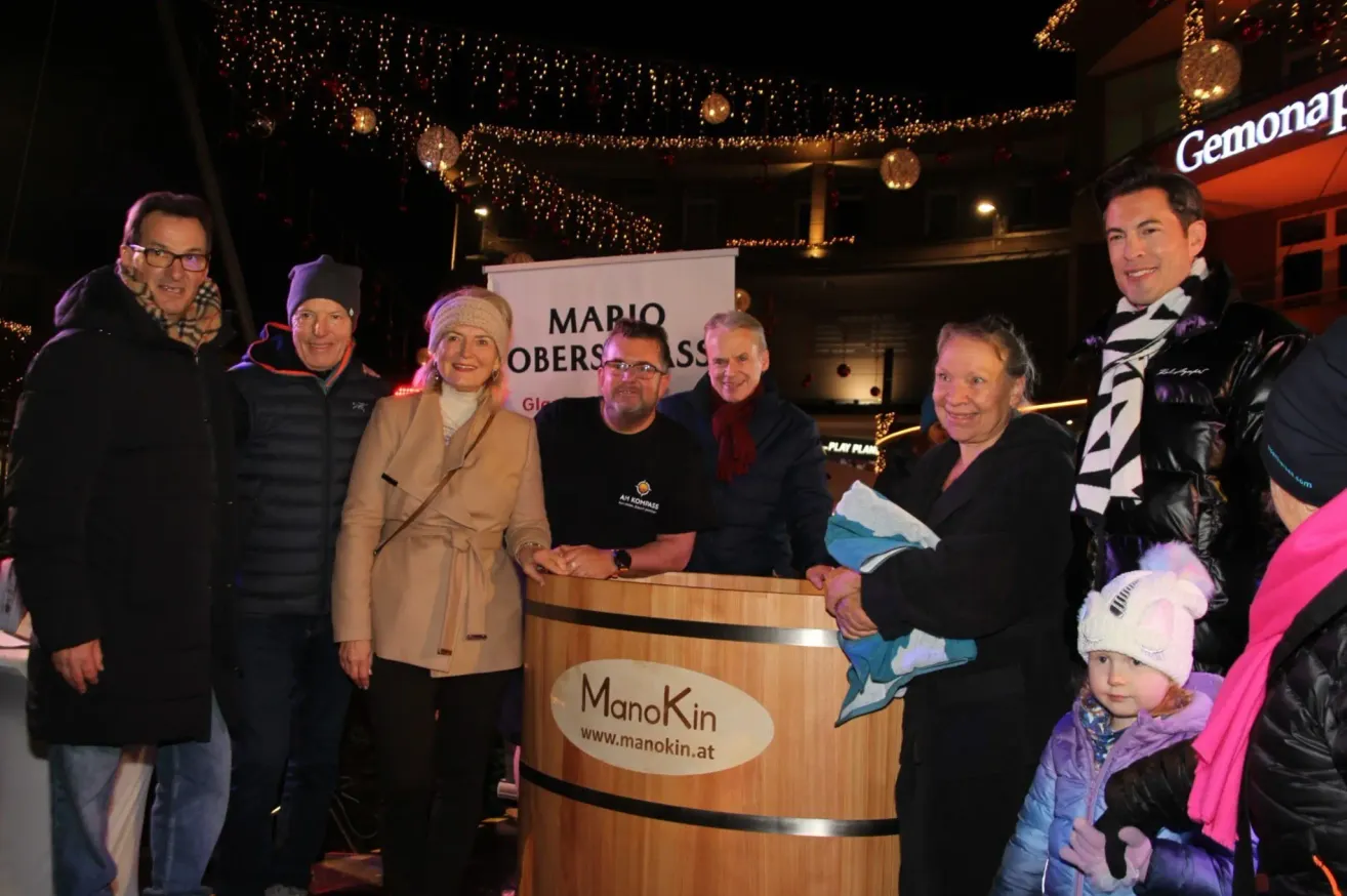 Eine Gruppe von Erwachsenen und einem Kind steht hinter einem hölzernen Fass mit dem Text ManoKin und einer Website. Sie lächeln und scheinen für ein Foto zu posieren. Dahinter steht ein Schild mit dem Text Mario Obers.