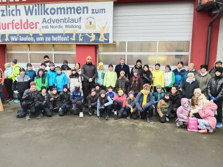 Eine Gruppe von Menschen in Winterkleidung steht vor einem Gebäude mit einem Banner, auf dem 'Willkommen zum Adventlauf mit Nordic Walking' steht. Sie posieren für ein Foto.