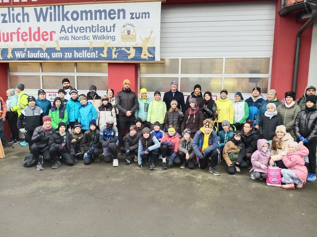 Eine Gruppe von Menschen in Winterkleidung steht vor einem Gebäude mit einem Banner, auf dem 'Willkommen zum Adventlauf mit Nordic Walking' steht. Sie posieren für ein Foto.