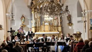 Ein Chor und Orchester spielen in einer Kirche mit goldenem Altar und Kronleuchter. Die Mitglieder sitzen auf der Bühne und halten Gitarren. Das Publikum sitzt auf den Kirchenbänken.