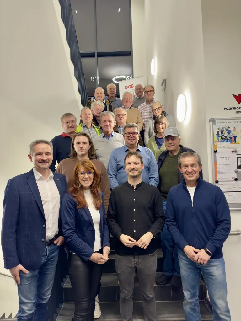 Eine Gruppe von Menschen, sowohl Männer als auch Frauen, steht mit lächelndem Gesichtsausdruck auf einer Treppe. Sie sind alle formell gekleidet. Ein rotes Pfeilschild und eine weiße Tafel mit Text sind an der Wand befestigt. Eine Lampe ist an der Wand auf der rechten Seite angebracht.