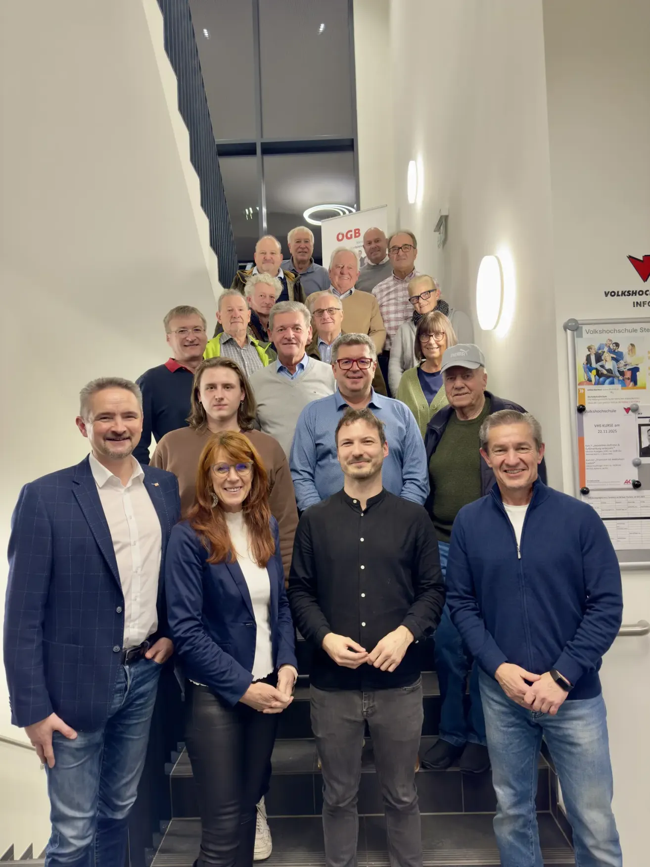 Eine Gruppe von Menschen, sowohl Männer als auch Frauen, steht mit lächelndem Gesichtsausdruck auf einer Treppe. Sie sind alle formell gekleidet. Ein rotes Pfeilschild und eine weiße Tafel mit Text sind an der Wand befestigt. Eine Lampe ist an der Wand auf der rechten Seite angebracht.