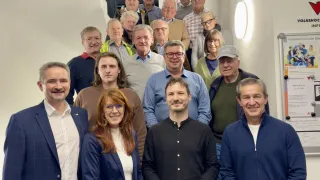 Eine Gruppe von Menschen, sowohl Männer als auch Frauen, steht mit lächelndem Gesichtsausdruck auf einer Treppe. Sie sind alle formell gekleidet. Ein rotes Pfeilschild und eine weiße Tafel mit Text sind an der Wand befestigt. Eine Lampe ist an der Wand auf der rechten Seite angebracht.