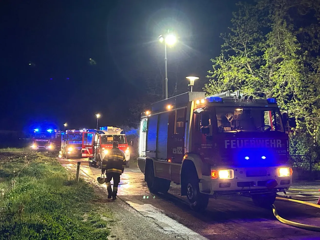 Feuerwehrleute in Schutzkleidung gehen bei Nacht auf einer nassen Straße. Mehrere Feuerwehrwagen mit eingeschalteten Lichtern geparkt.