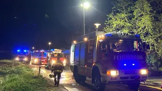 Feuerwehrleute in Schutzkleidung gehen bei Nacht auf einer nassen Straße. Mehrere Feuerwehrwagen mit eingeschalteten Lichtern geparkt.